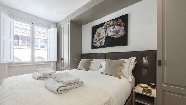A bedroom with a large bed, a nightstand, a wall-mounted painting, and a window with open shutters.