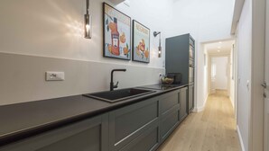 A modern kitchen with a dark countertop, a built-in sink, and a backsplash. There are two framed pictures on the wall, a light fixture above the sink, and a cabinet with a microwave.