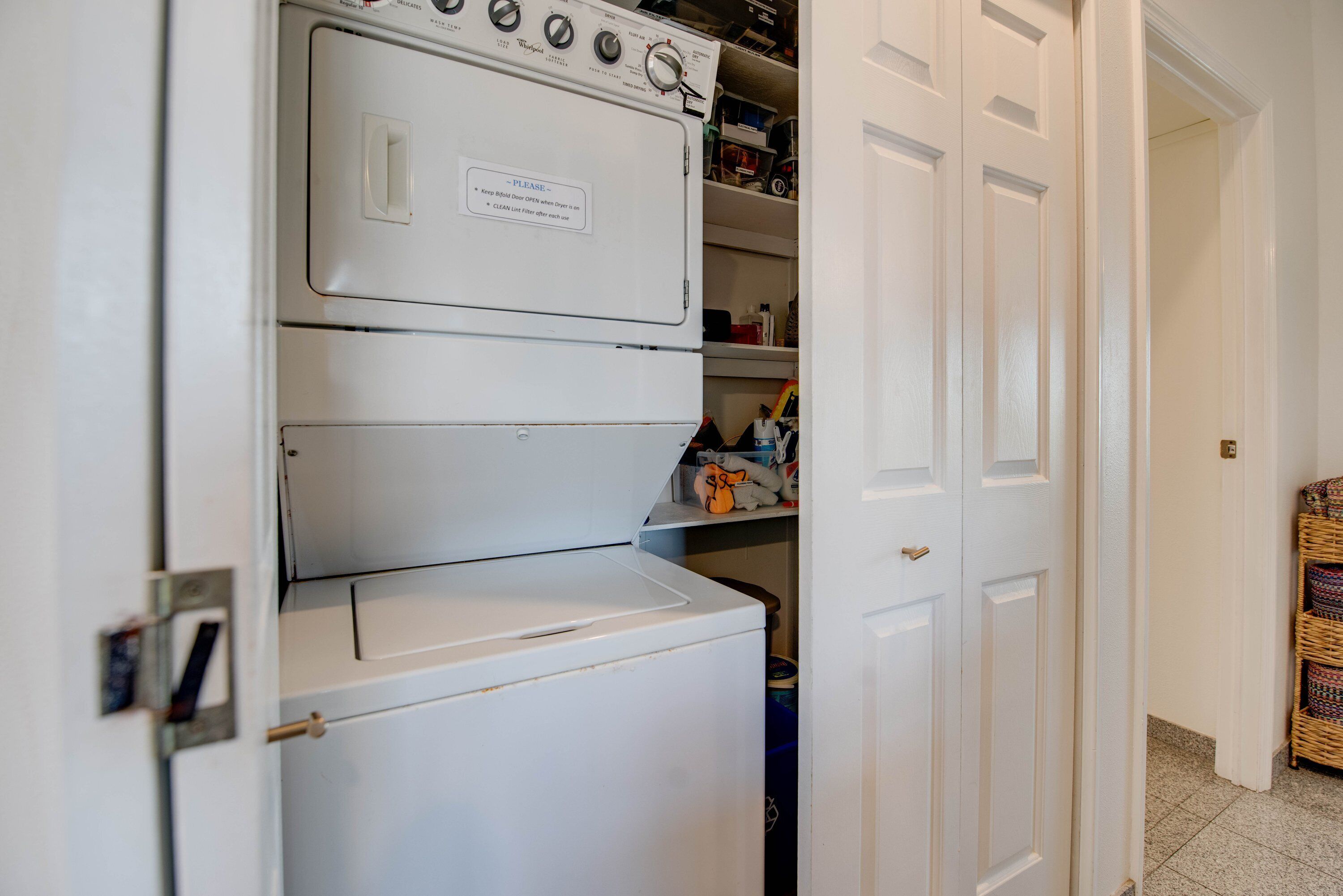 A laundry room with stacked washers and dryers, a shelf with cleaning supplies, and a door leading to another room.