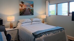 Une chambre à coucher avec un lit, deux tables de chevet avec des lampes, un ventilateur de plafond, une fenêtre avec des stores et un tableau représentant une scène de plage.