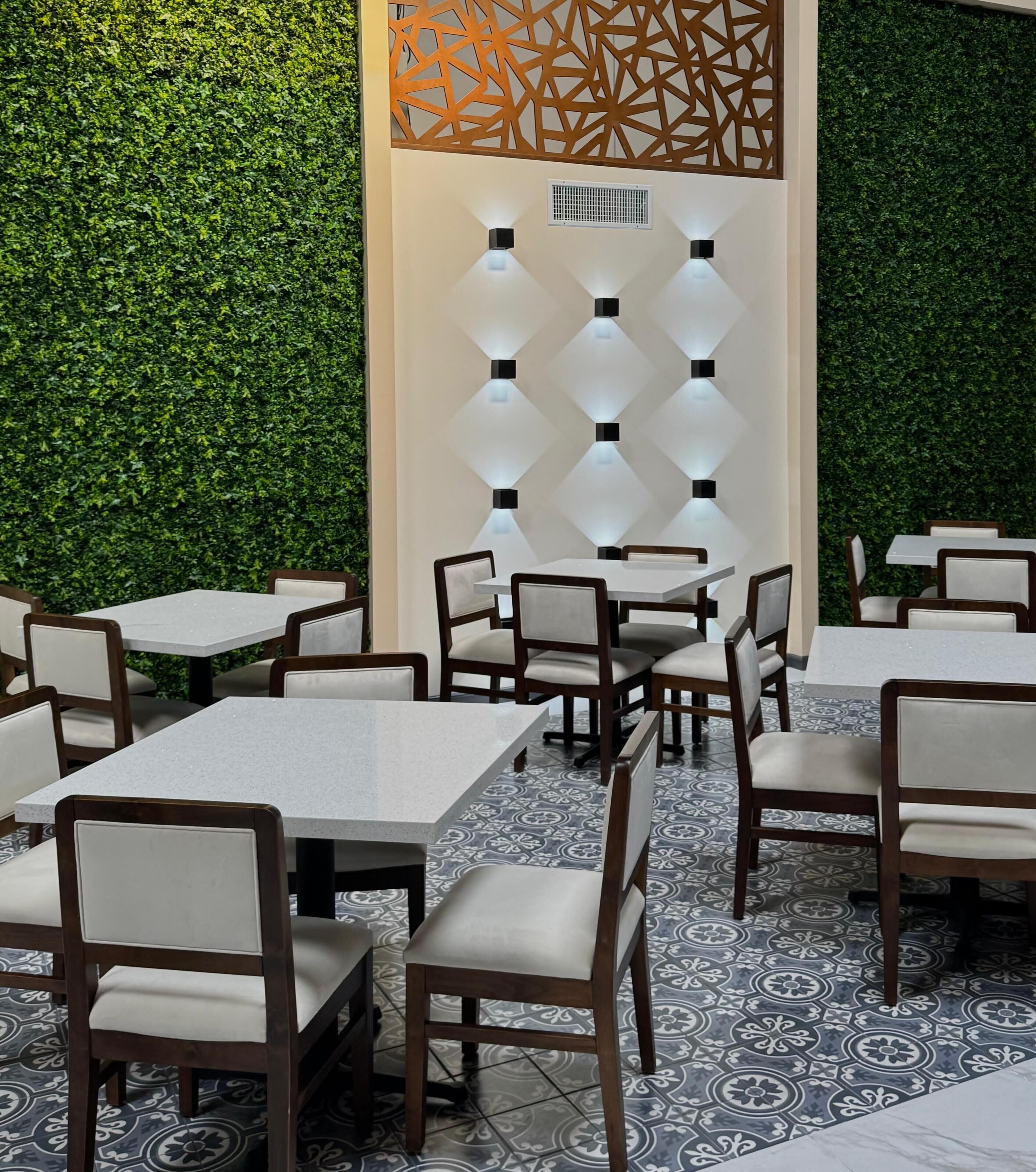 An outdoor dining area with tables and chairs, a decorative wall with geometric patterns, and a green hedge backdrop.