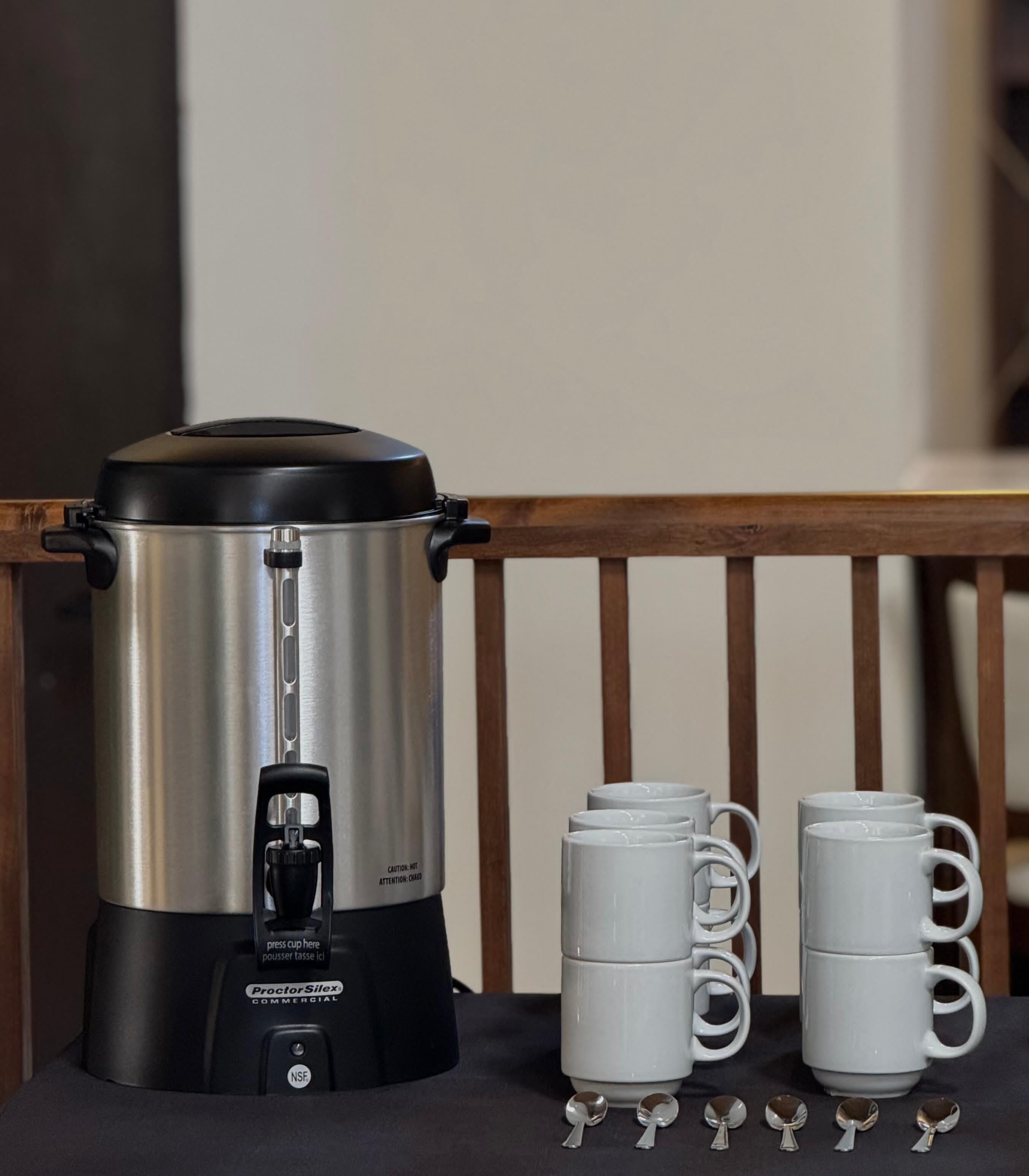 Une cafetière professionnelle ProctorSilex en acier inoxydable, dotée d’un couvercle noir, d’un piston à café et de quatre tasses blanches à anse, posées sur une table.