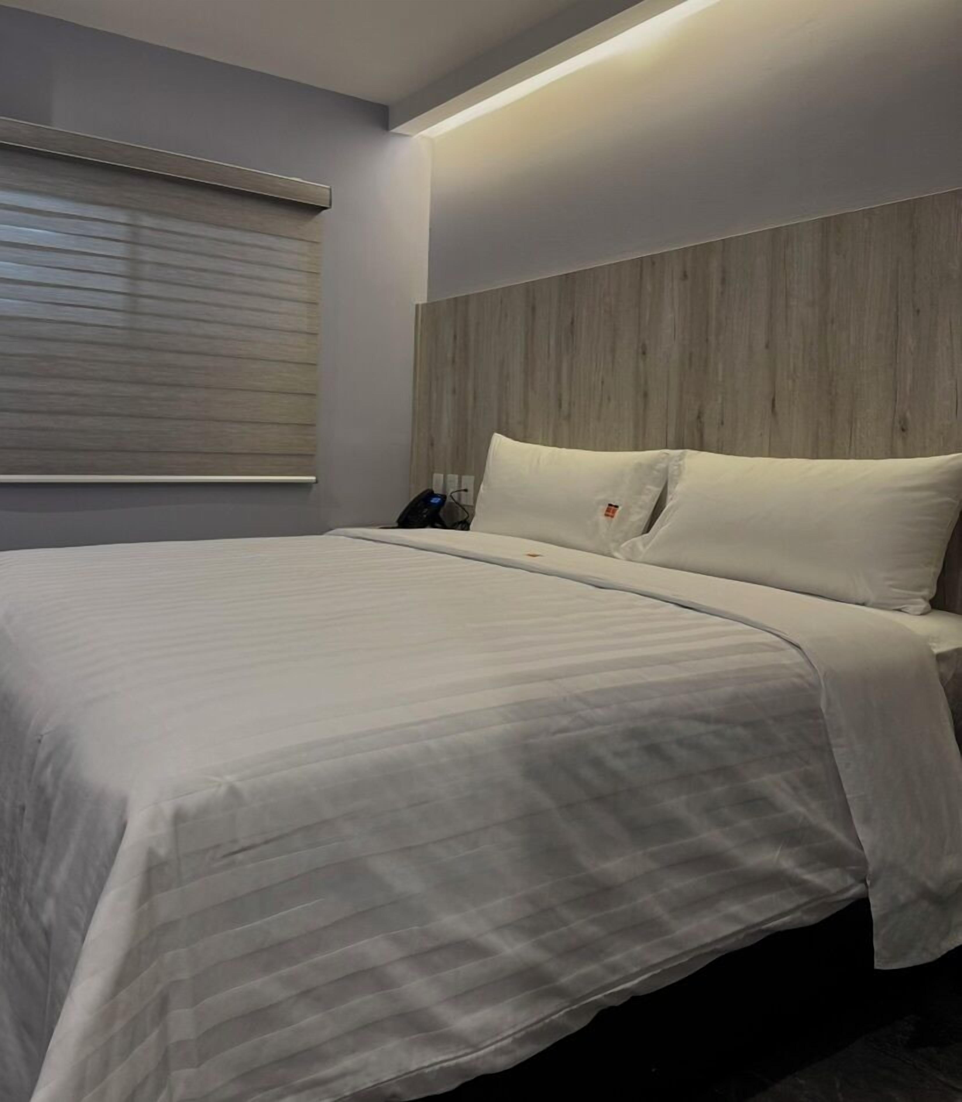 A neatly made bed with a wooden headboard, a bedside phone, and a window with blinds.