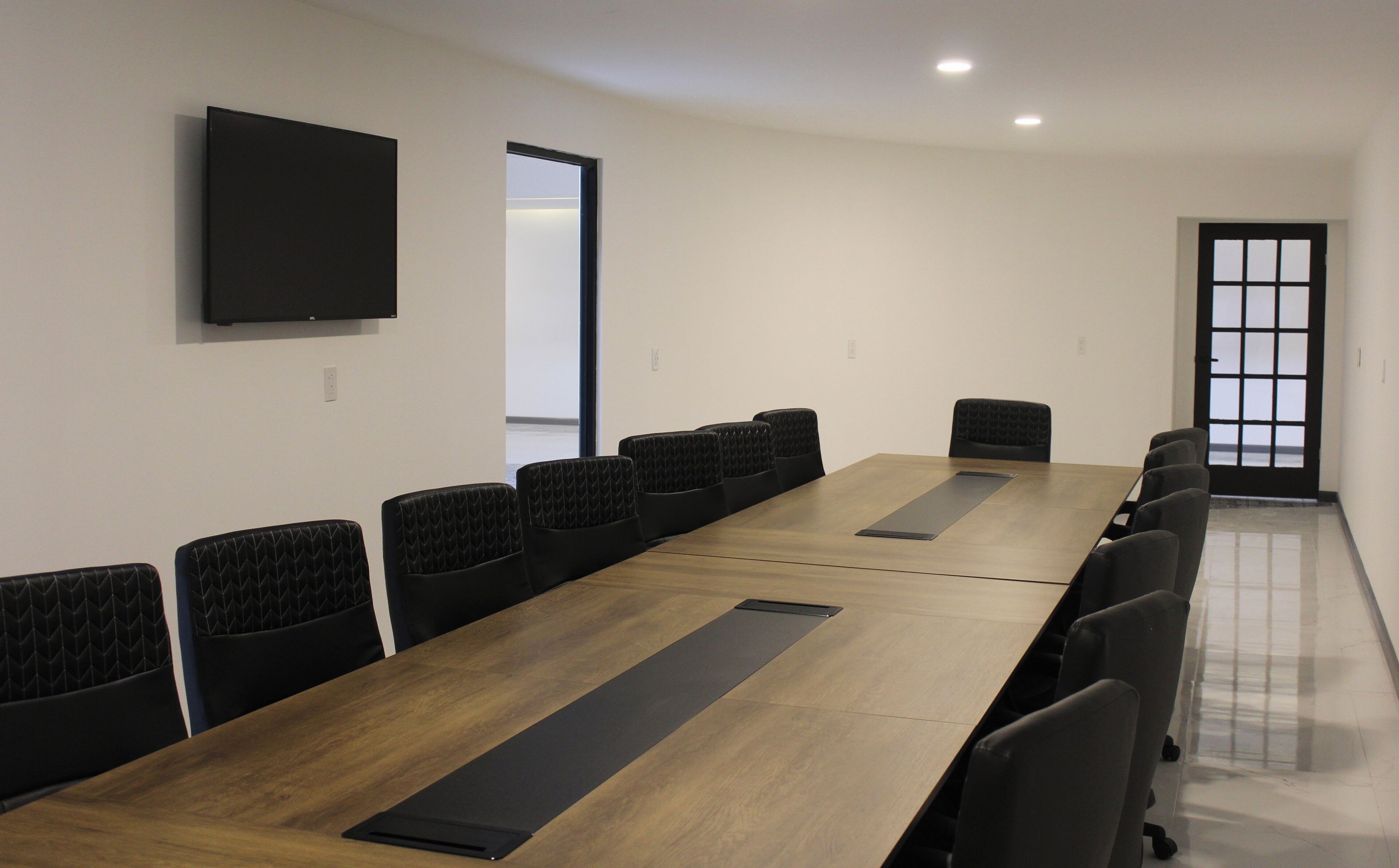 A conference room with a long table, black upholstered chairs, a flat-screen TV mounted on the wall, and a door with a window view.