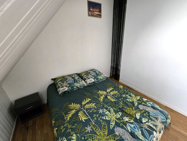 A small, neatly arranged bedroom with a bed, two pillows, a nightstand, and a framed picture on the wall.