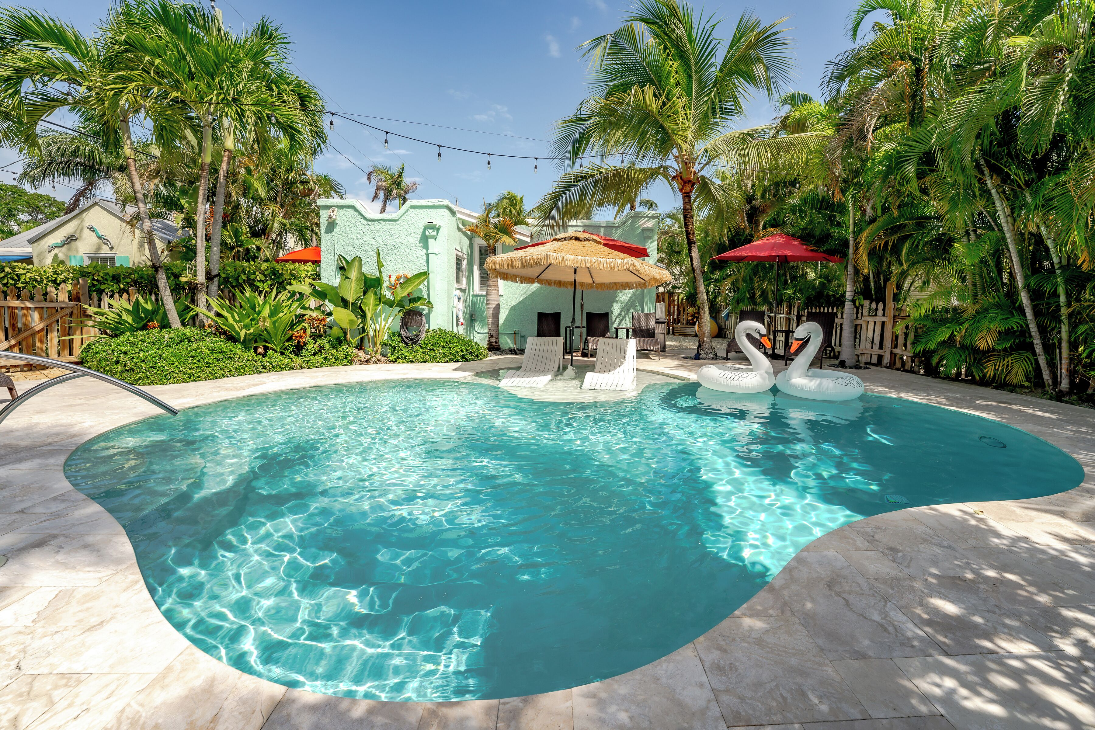 A swimming pool with a curved edge, surrounded by palm trees and a small gazebo with a red umbrella.
