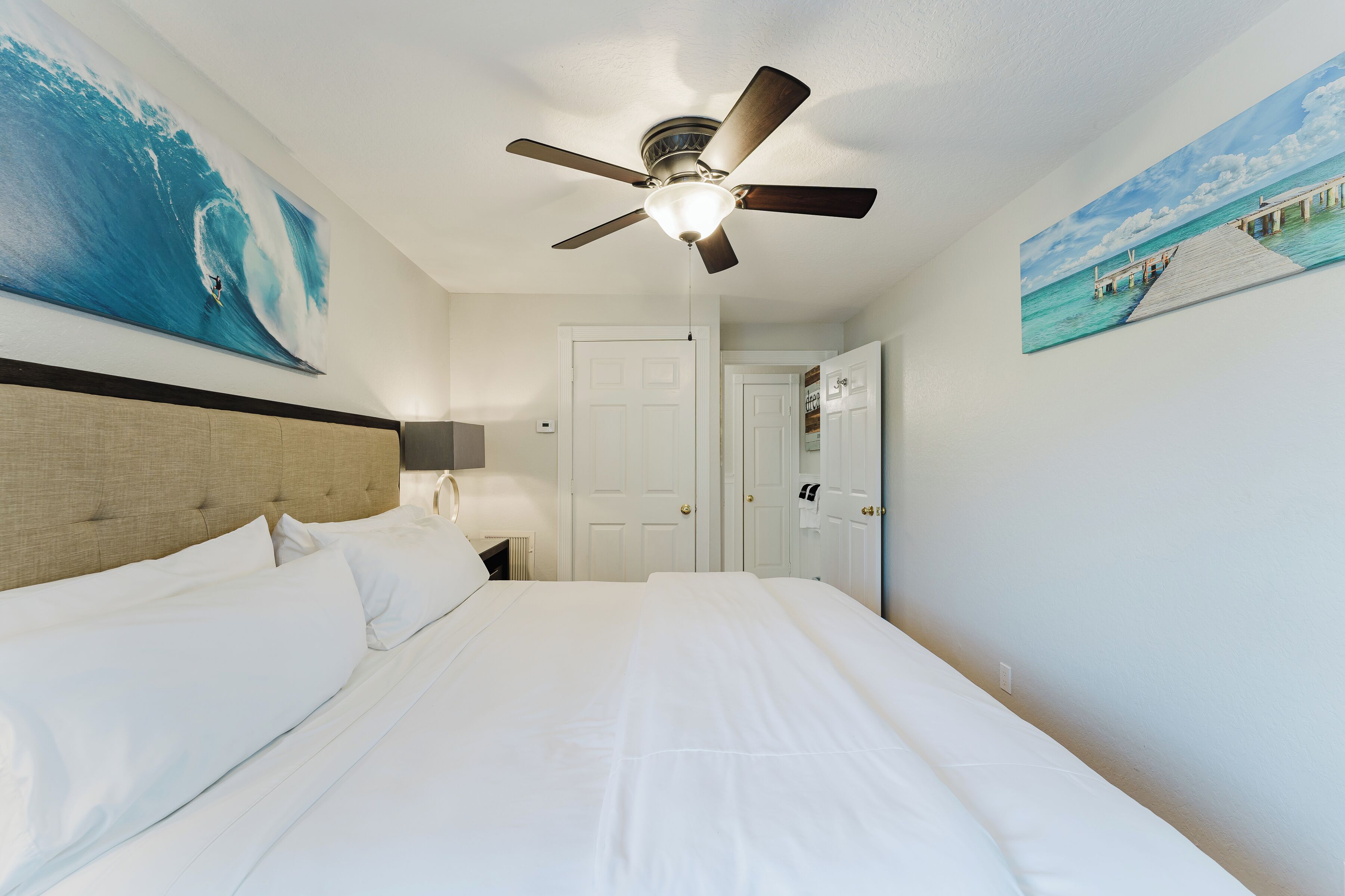 Habitación de hotel con una cama, un ventilador de techo, un cuadro de un surfista en una ola y un cuadro de un muelle.