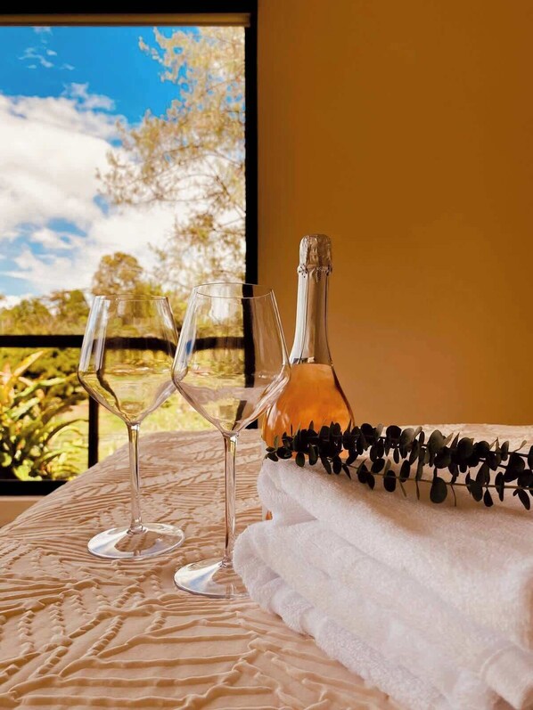 Una mesa con dos copas de vino y una botella de champán, sobre un mantel texturizado, con vistas a árboles y al cielo a través de una ventana.