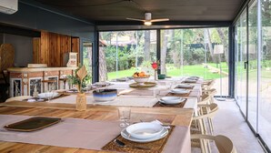 A dining area with a long table set for a meal, large windows offering a view of the outdoors, and a ceiling fan overhead.