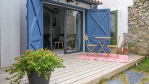 Une terrasse en bois agrémentée d’un pot de fleurs, d’une couverture à carreaux rouges et blancs, et d’une table pliante avec des chaises.