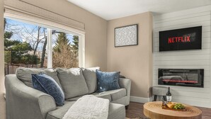 A living room with a grey sofa, a white throw blanket, a round wooden coffee table with a bottle and snacks, a mounted TV displaying 'NETFLIX', and a framed picture on the wall. - LUX Guest Suite Near Amazing Hiking Trails (Colorado Springs)