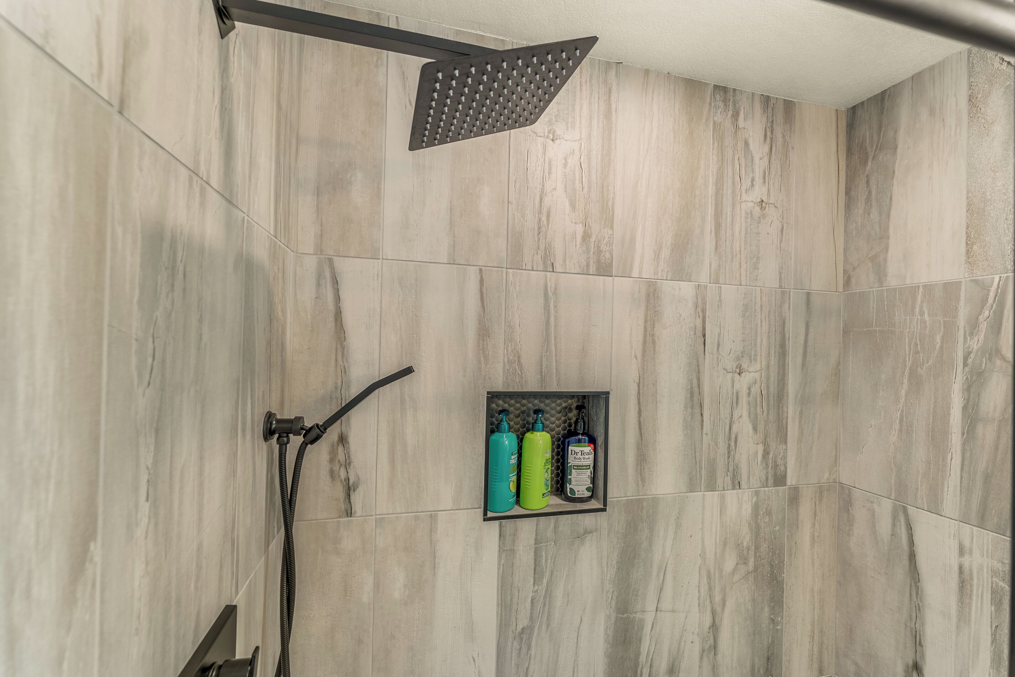 A shower area with a rain showerhead, a handheld showerhead, and a shelf with toiletries.