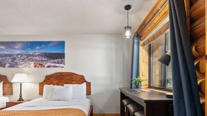 Habitación de hotel con una cama de madera, un escritorio con sillas, una ventana con cortinas y un cuadro en la pared.