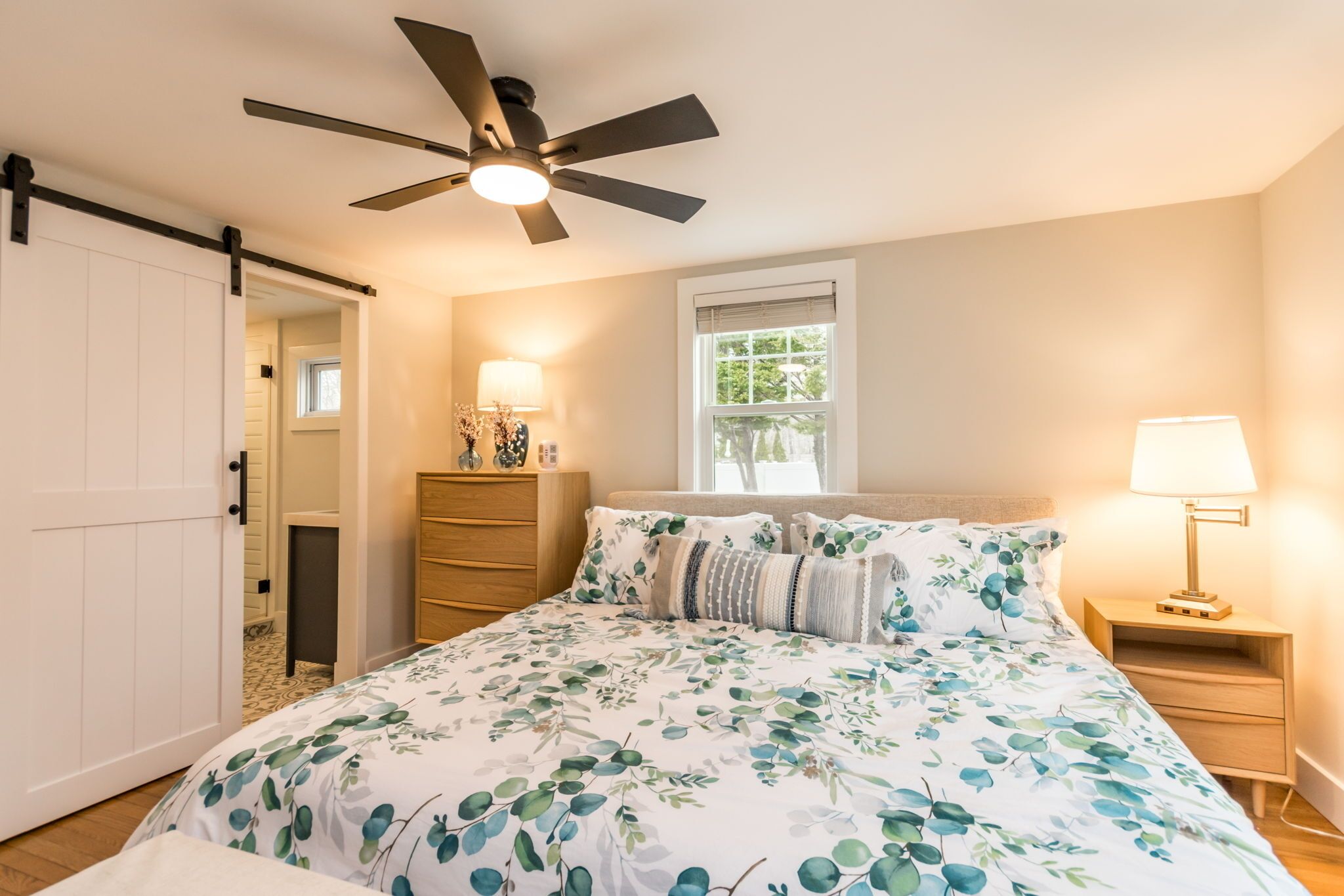 A bedroom with a bed, a ceiling fan, a nightstand, a lamp, a window, and a door.