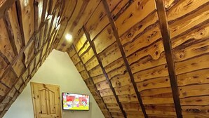 A bedroom with a sloped ceiling, wooden beams, a bed with white linens, a TV on the wall, and a wooden door.