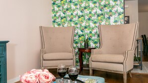A room with two beige armchairs, a small wooden table with a vase of pink flowers, and two wine glasses.