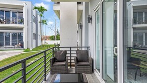 Ein Balkon mit Korbmöbeln und einem Glastisch, mit Blick auf eine gepflegte Rasenfläche und angrenzende Gebäude.