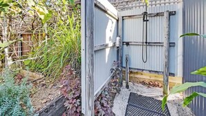 Une porte en bois mène à un espace extérieur abrité, doté d’un toit en métal, et cet espace est entouré de végétation et d’une clôture en bois.