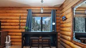 A log cabin interior with a wooden dining table, two chairs, a potted plant, and a view of trees through a window.