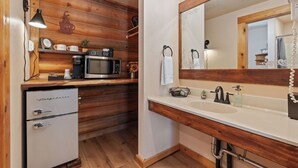 A compact kitchen with wooden paneling, a coffee maker, a microwave, a refrigerator, and a sink with a mirror above it.
