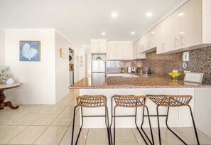 A modern kitchen with a granite countertop, white cabinets, and a built-in oven. There are three wicker bar stools and a small dining table with a vase.