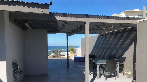 A covered outdoor area with a table and chairs, a view of the ocean, and a building with a corrugated metal roof.