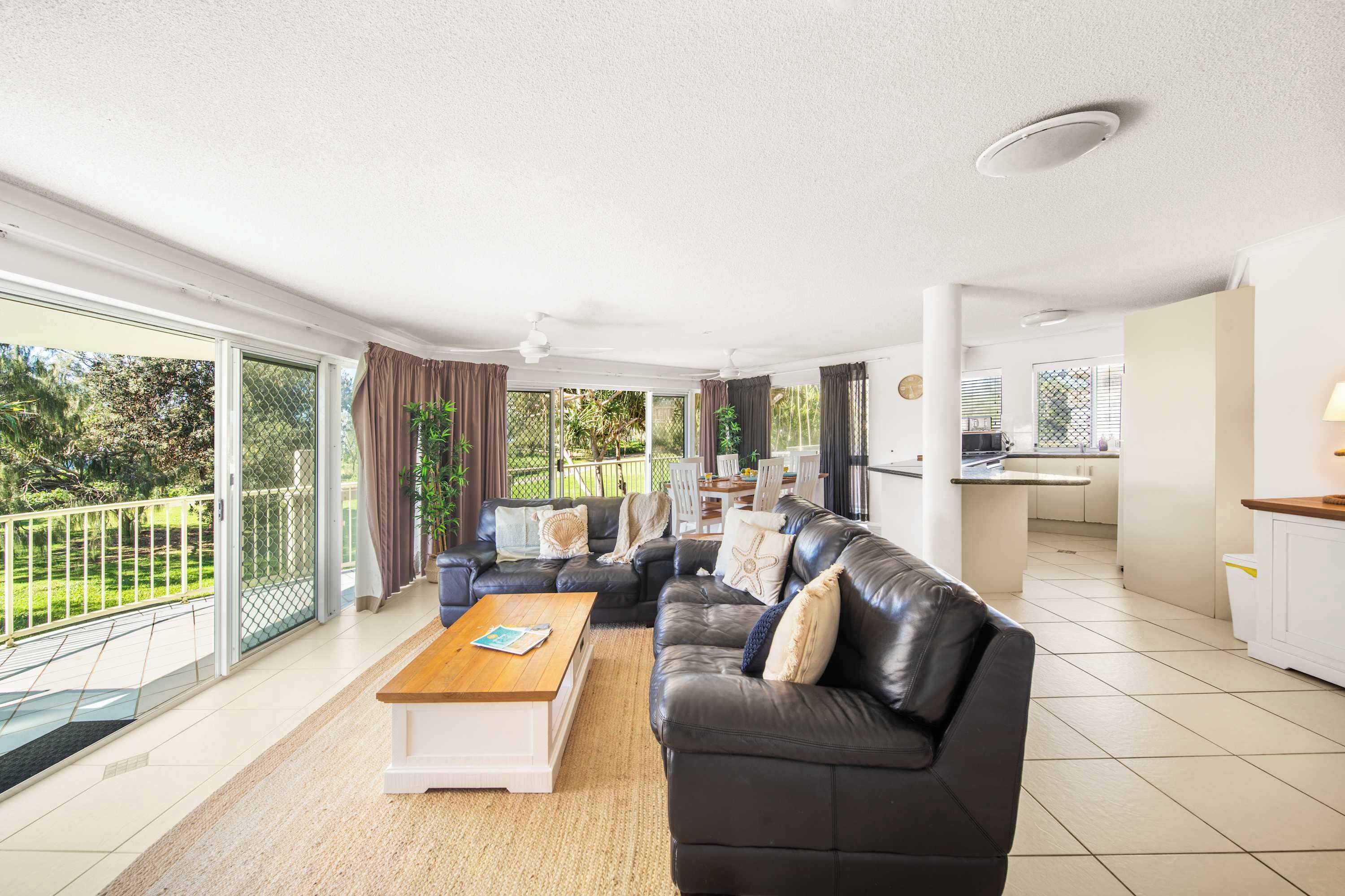 A spacious living area with a black leather sofa, a wooden coffee table, and a view of a garden through large sliding glass doors.