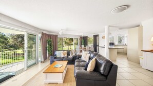 A spacious living area with a black leather sofa, a wooden coffee table, and a view of a garden through large sliding glass doors. - Surfside Beachfront 3bed Apt - Direct Beach Access (Buddina)