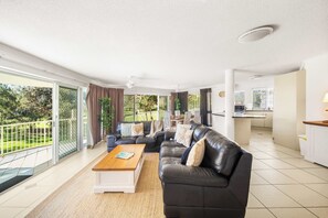 A spacious living area with a black leather sofa, a wooden coffee table, and a view of a garden through large sliding glass doors.