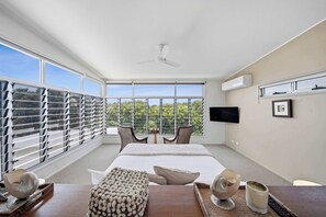 A modern hotel room with a large bed, a ceiling fan, a TV mounted on the wall, and a balcony with a view of the trees.
