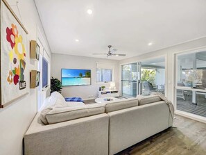 Ein modernes Wohnzimmer mit einer großen Couch, einem Flachbildfernseher, einem Deckenventilator und Blick ins Freie durch große Glasüren.