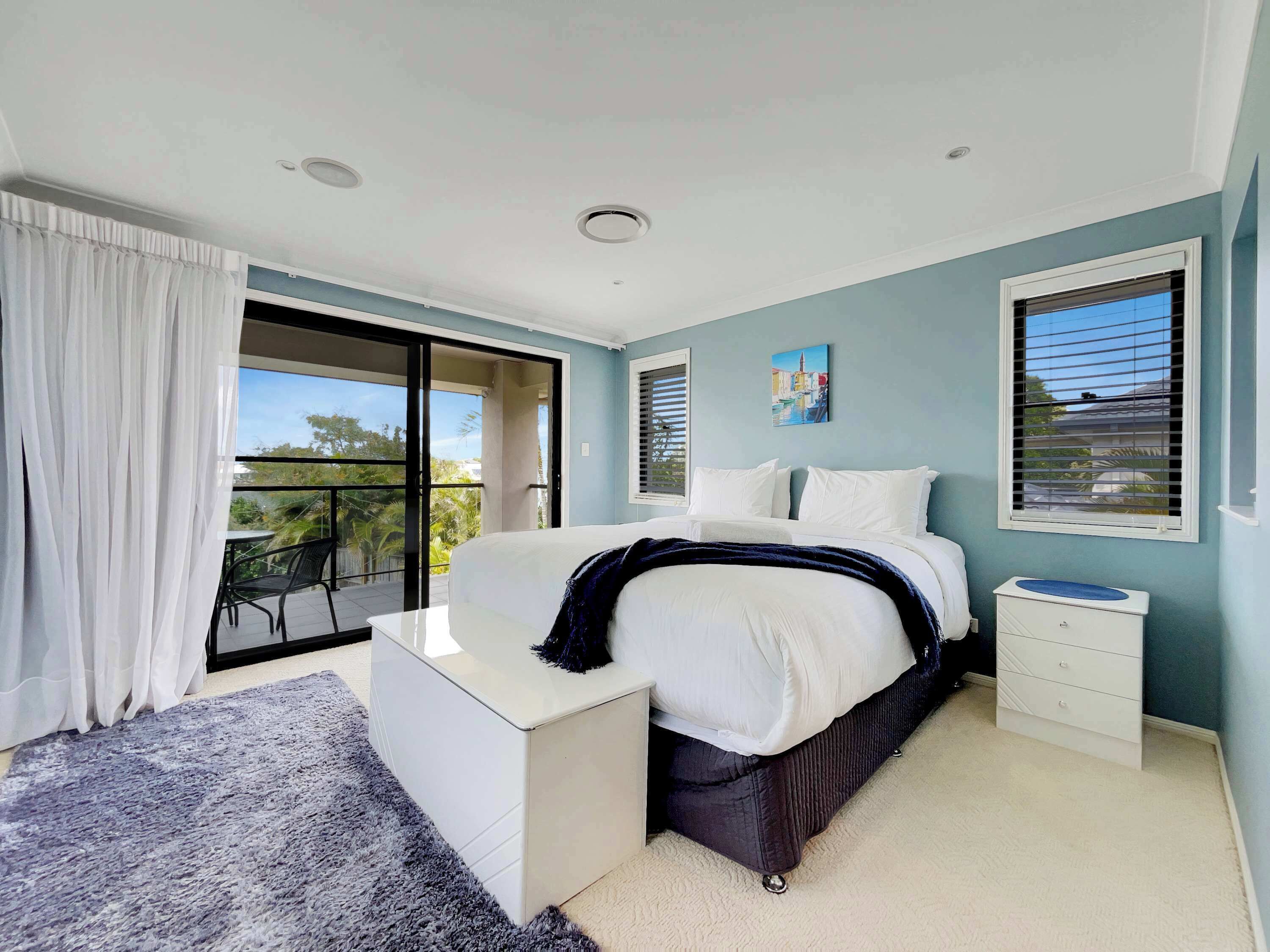 A bedroom with a bed, a nightstand, a window with blinds, a painting, and a balcony with a chair.