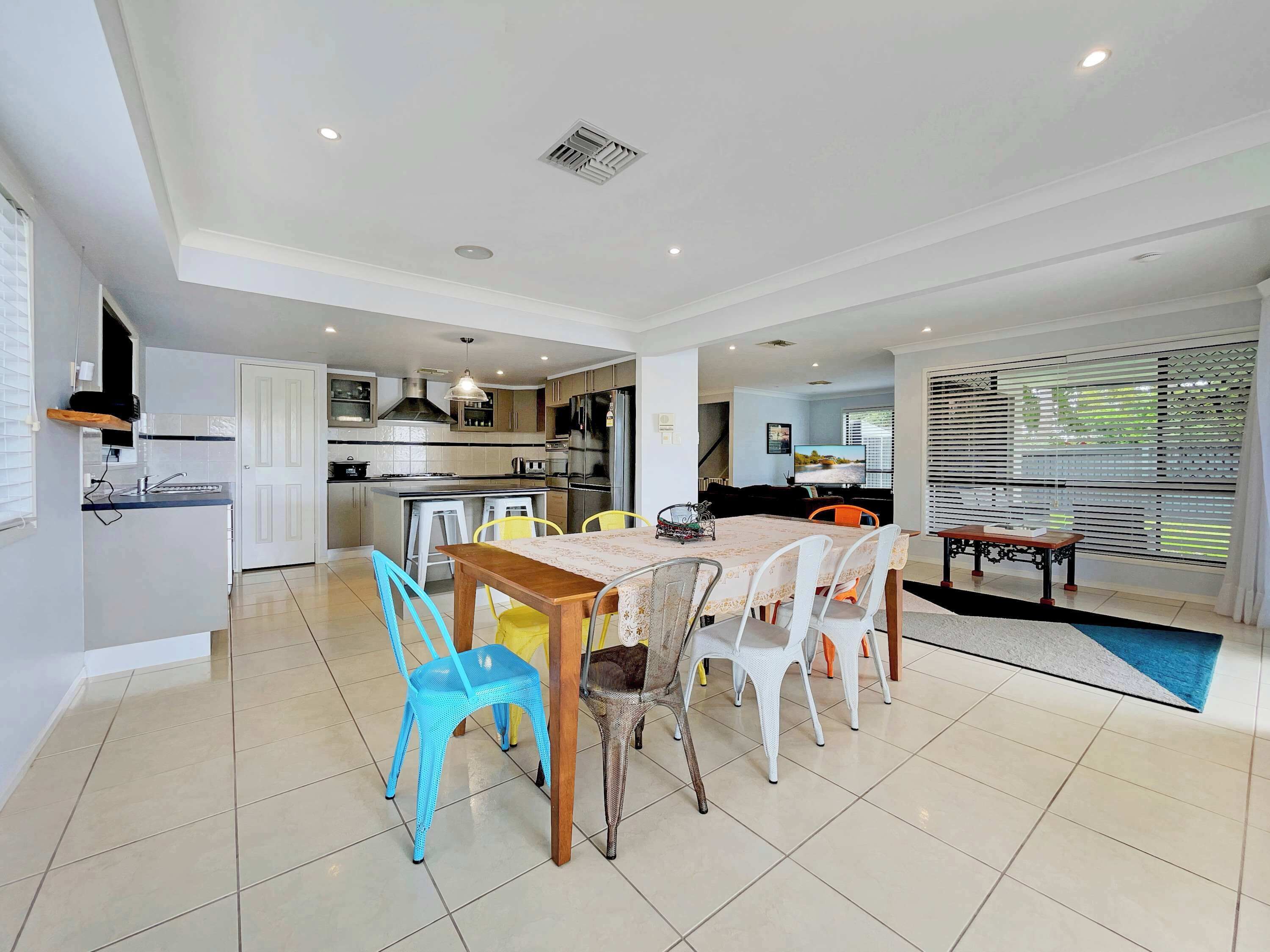 A modern kitchen with white cabinets, a dining area with a wooden table and chairs, and a living room with a TV and blinds.