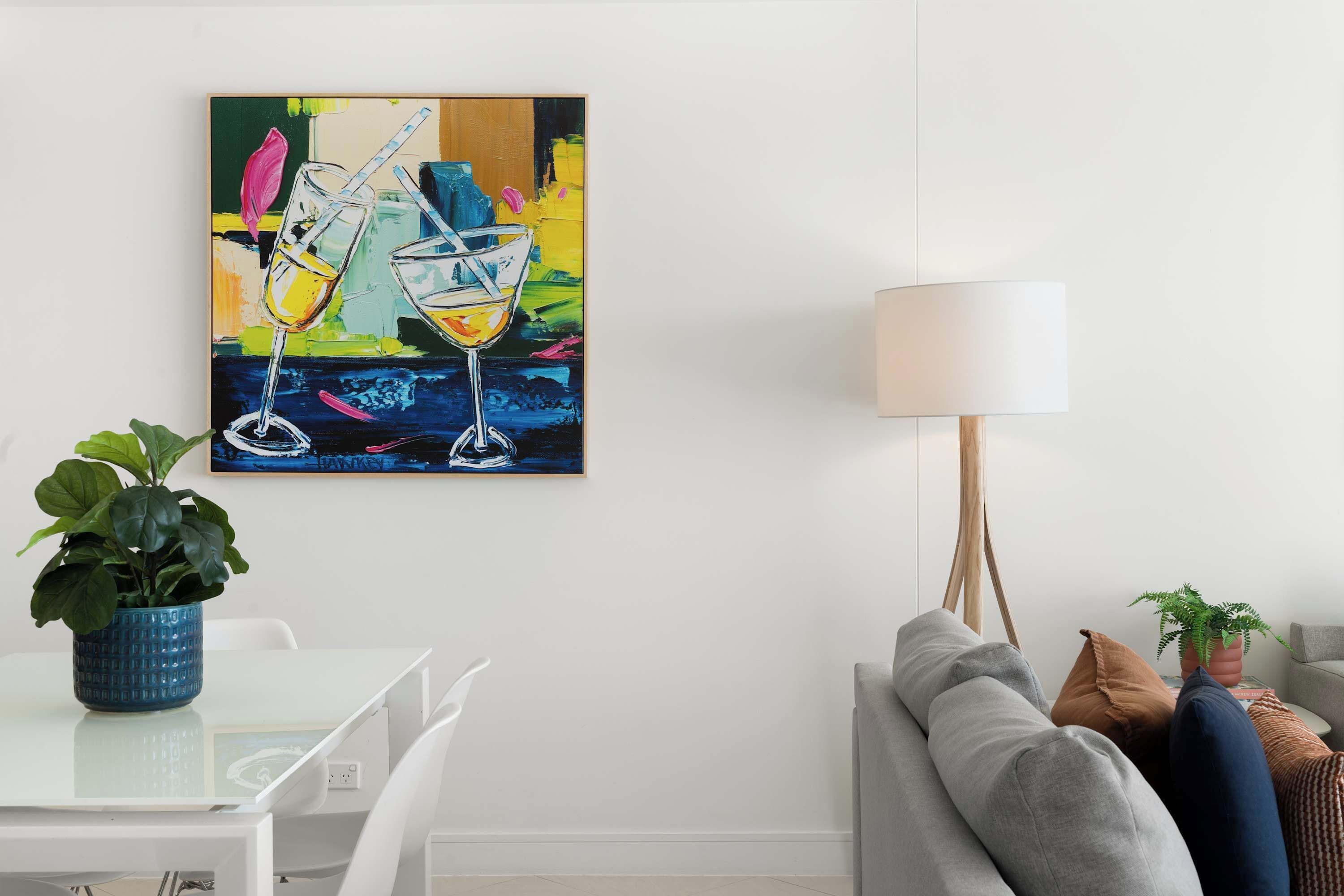Uma sala de estar moderna com uma mesa branca, um sofá cinzento, um vaso azul com planta e um quadro abstrato colorido na parede.