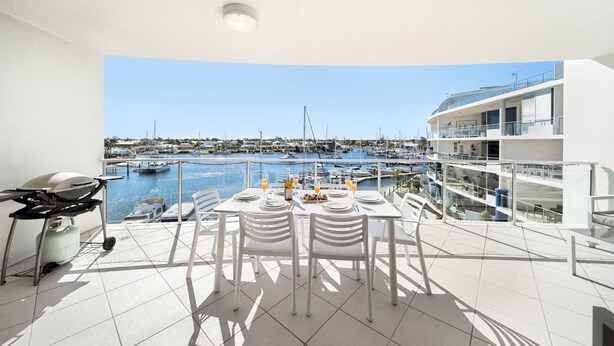 A balcony with a dining table set for two, overlooking a marina with boats and buildings.