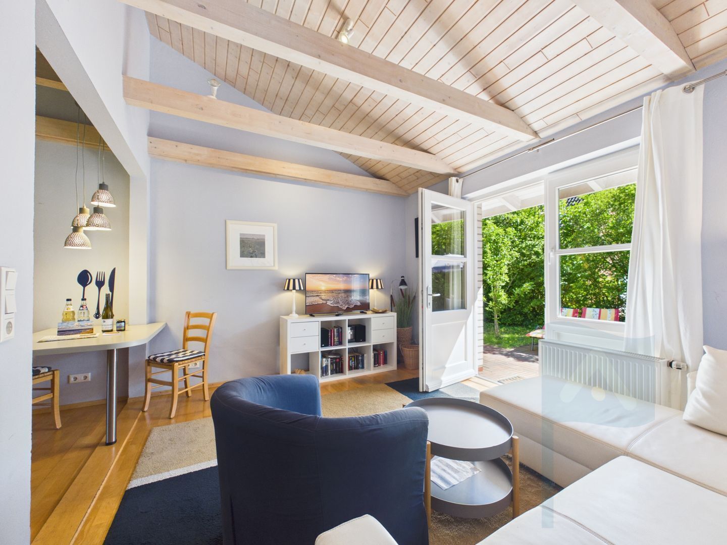 Un salon moderne avec un plafond en bois, un coin repas comprenant une table et des chaises, un canapé, un téléviseur et une fenêtre donnant sur un paysage verdoyant.