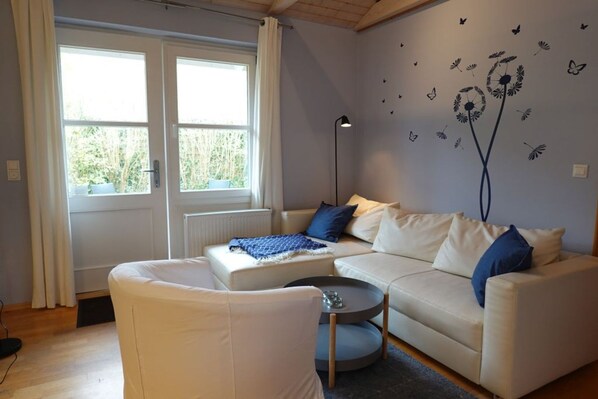 A modern living room with a white sofa, a small round table, and a floor lamp. There is a window with curtains, and the walls are decorated with butterfly decals.
