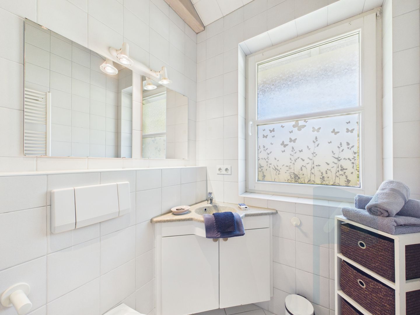 Een badkamer met een betegelde muur, een grote spiegel, een raam met vlinderdecoratie, en een handdoekhouder.