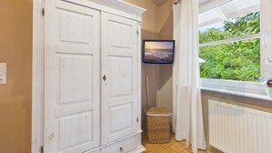 Een kamer met een witte garderobe, een aan de muur bevestigde televisie, een raam met gordijnen en een radiator.