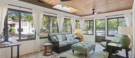 A sunroom with a herringbone floor, a wicker sofa, a matching ottoman, a side table, and a ceiling fan.