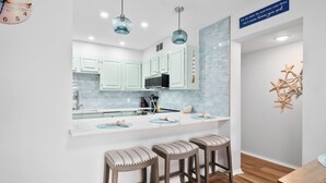 A modern kitchen with light green cabinets, a white countertop, and a central island with bar stools. There are pendant lights above the island and a clock on the wall.