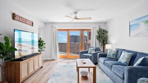 A living room with a large TV, a blue sofa, a wooden cabinet, and a view of the beach through the sliding glass doors.