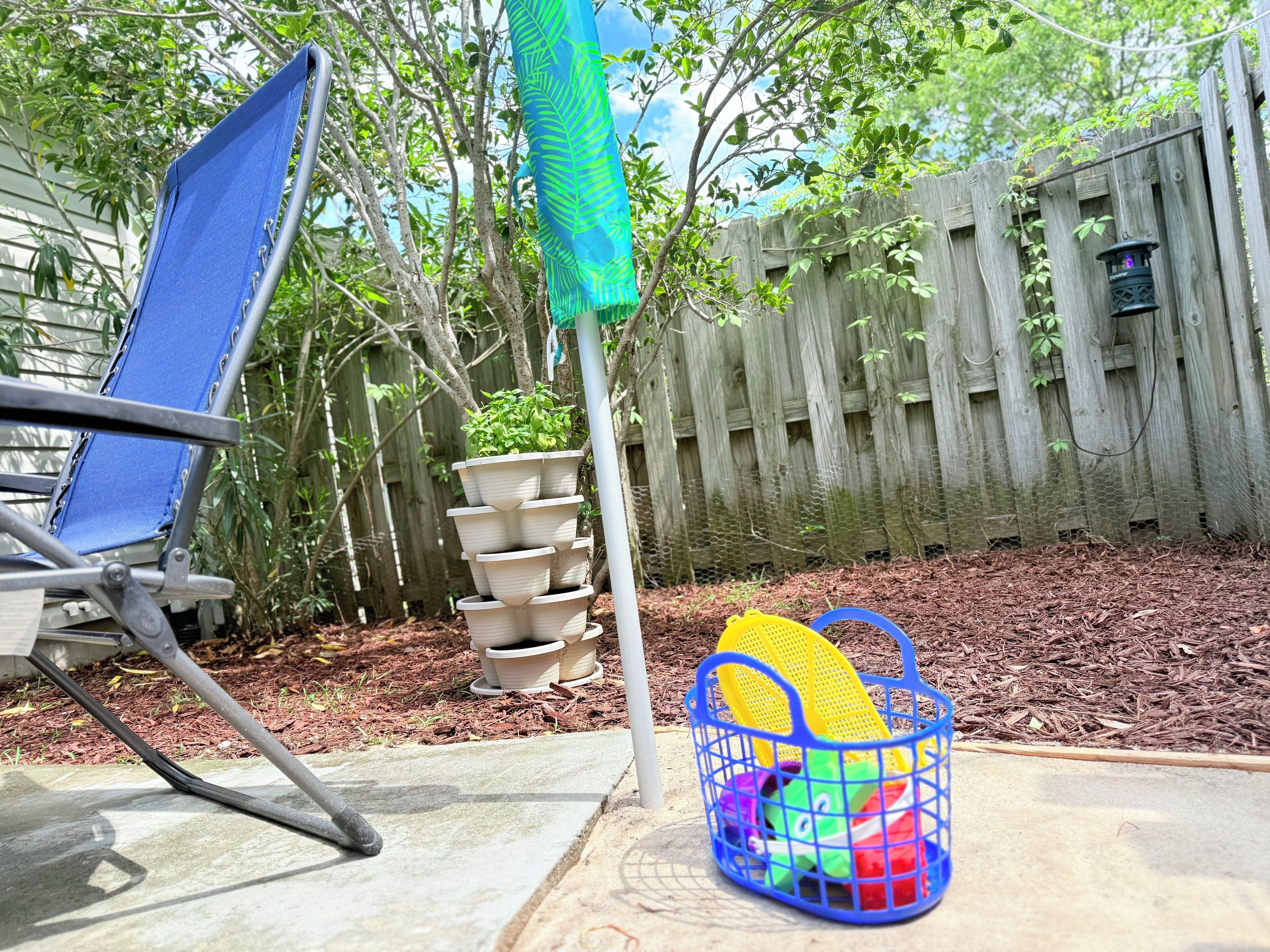 A backyard with a wooden fence, a blue chair, a stack of white pots with plants, and a blue basket with colorful balls.