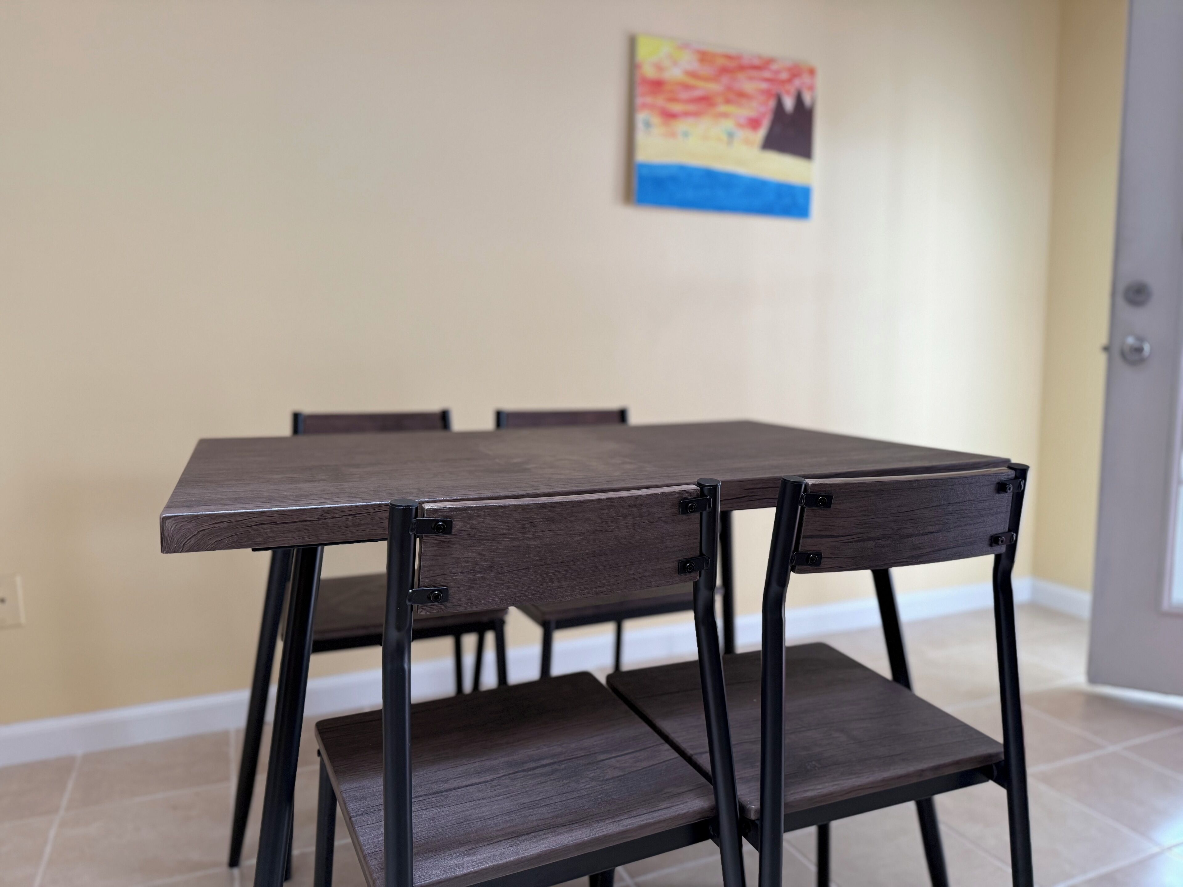 Uma mesa de jantar com quatro cadeiras, um quadro na parede e uma porta ao fundo.