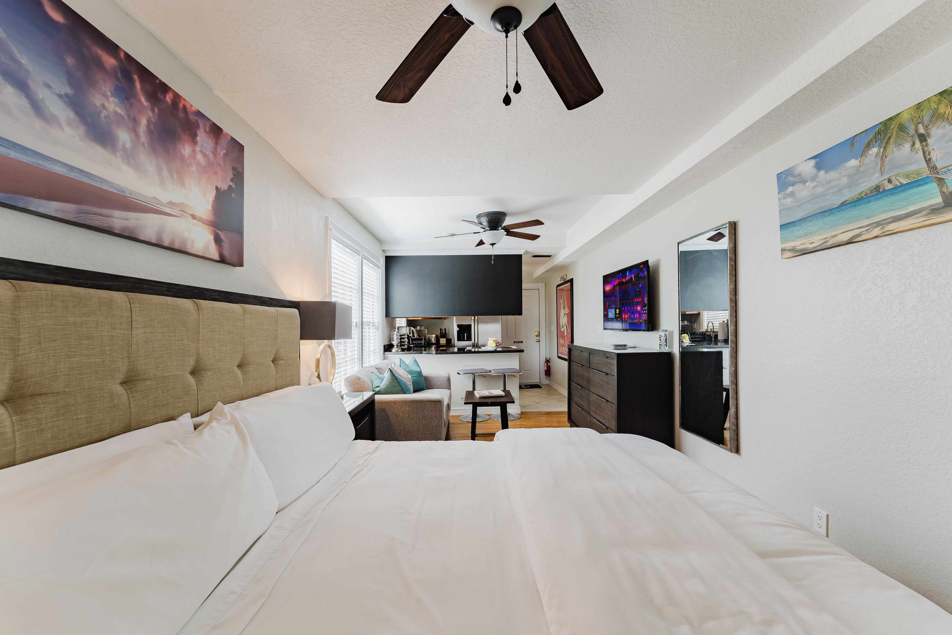 A modern hotel room with a large bed, a ceiling fan, a kitchenette, and artwork on the walls.