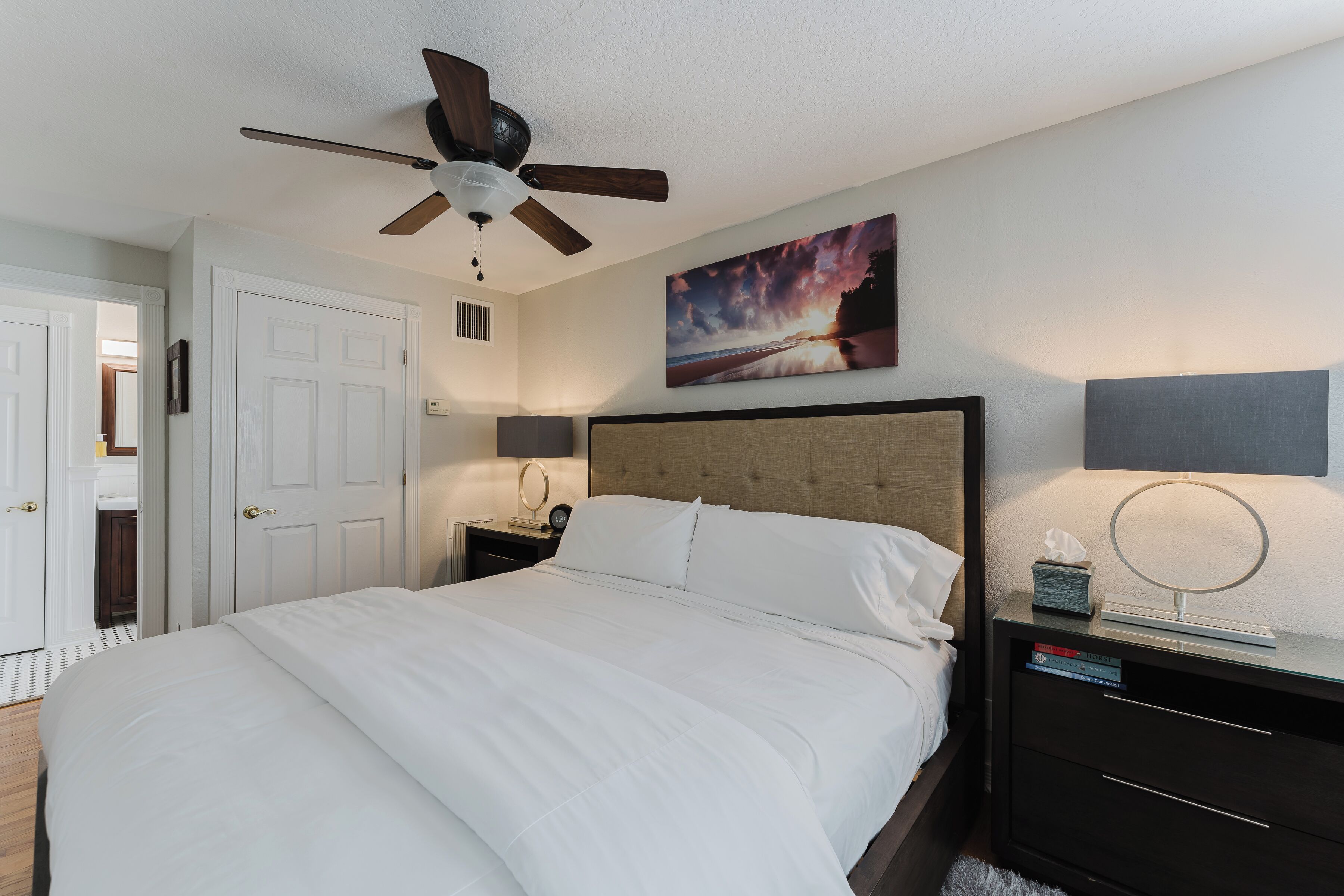 A neatly arranged bedroom with a large bed, a ceiling fan, a bedside table with a lamp, and a framed picture on the wall.