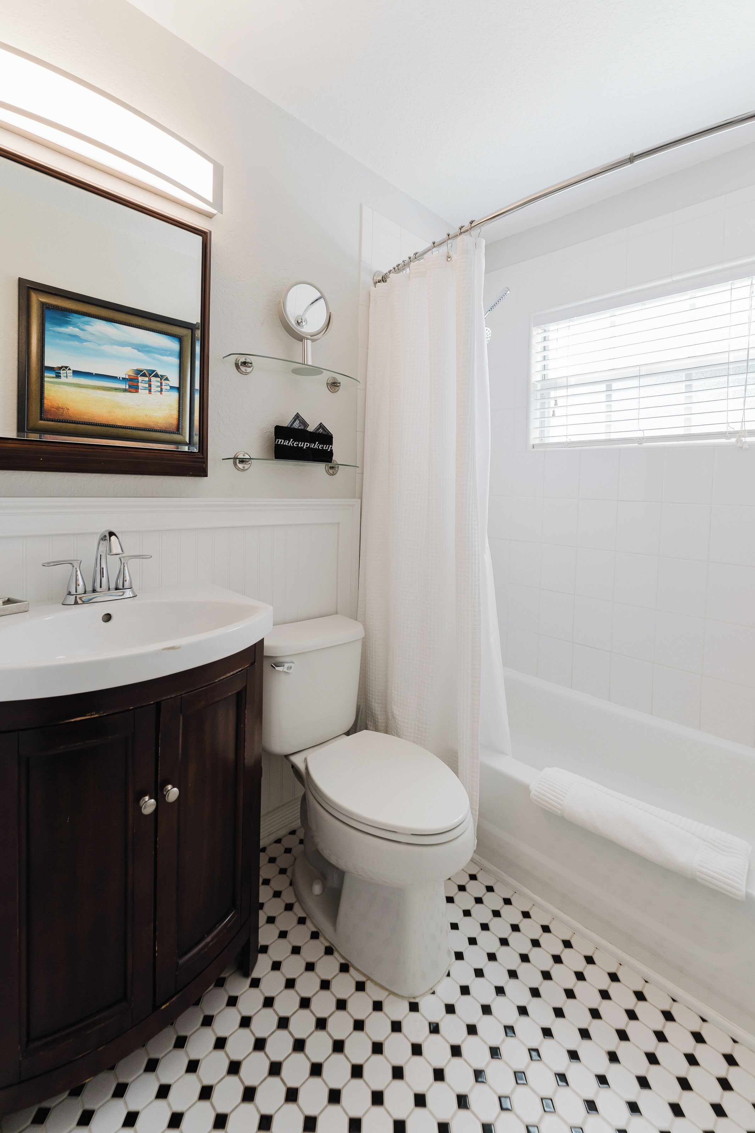 A bathroom with a white toilet, a dark wooden vanity, a white sink, a mirror, a framed picture on the wall, and a window with a white curtain.