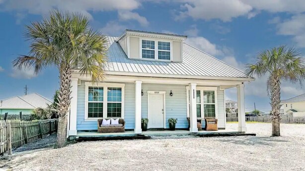 A coastal house with a covered porch, palm trees, and a clear sky.