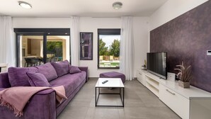 Una sala moderna con un sofá púrpura, una mesa de centro blanca, un televisor de pantalla plana y un amplio ventanal con vistas al exterior.