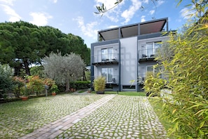 Un edificio moderno de dos pisos con un patio empedrado, plantas en macetas y un gran árbol al fondo.